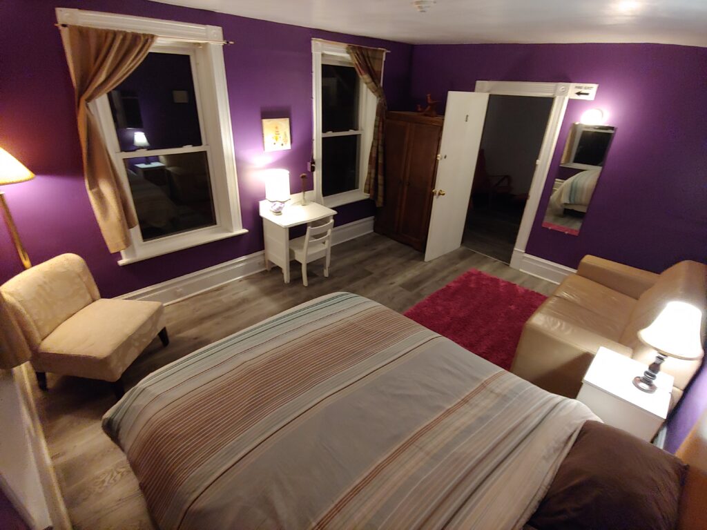 purple room at night with bed, pink rug, loveseat, chair two lamps, wardrobe