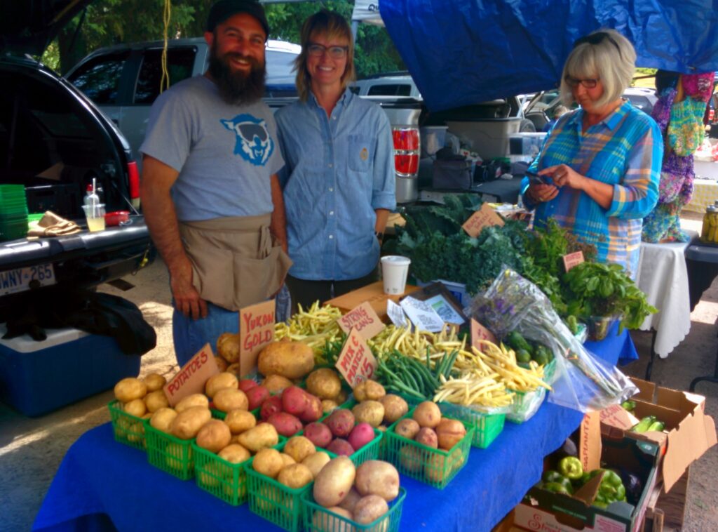 proprietors and customer behind table of vegetables at farmer's market