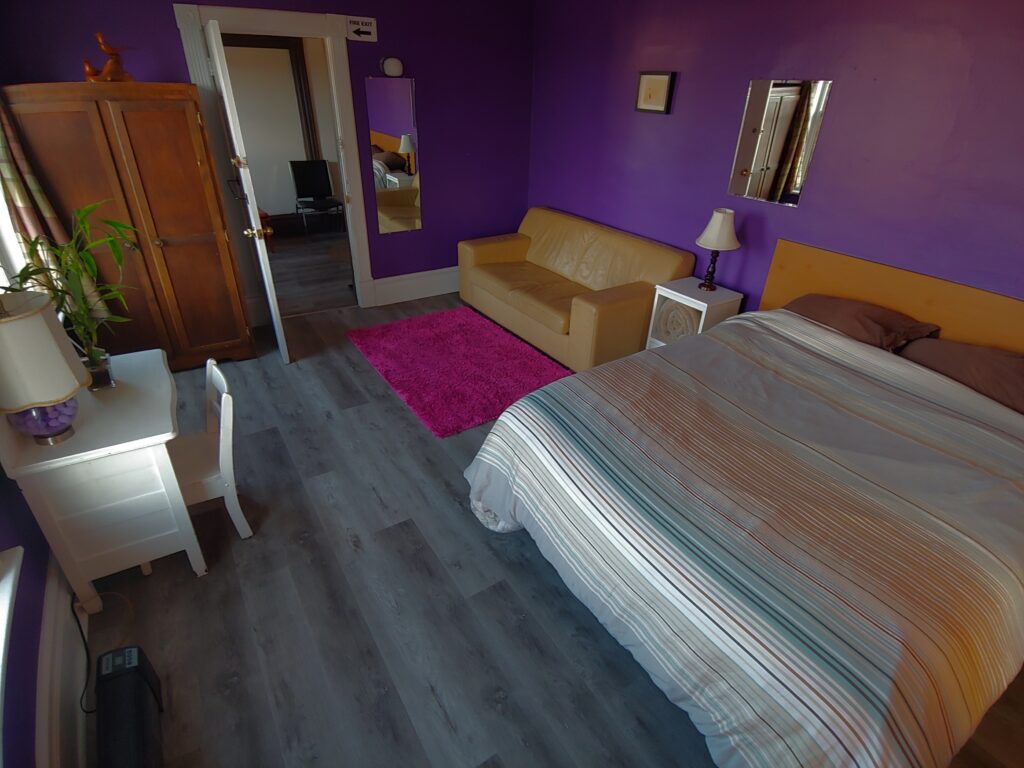 queen bed, couch, night table, two mirrors, wardrobe, tiny desk & chair, pink rug, purple walls