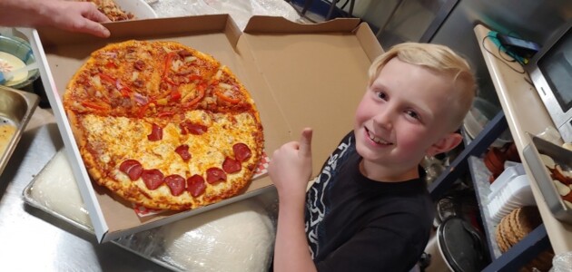 a kid giving the thumbs up beside a pizza with a smiley face